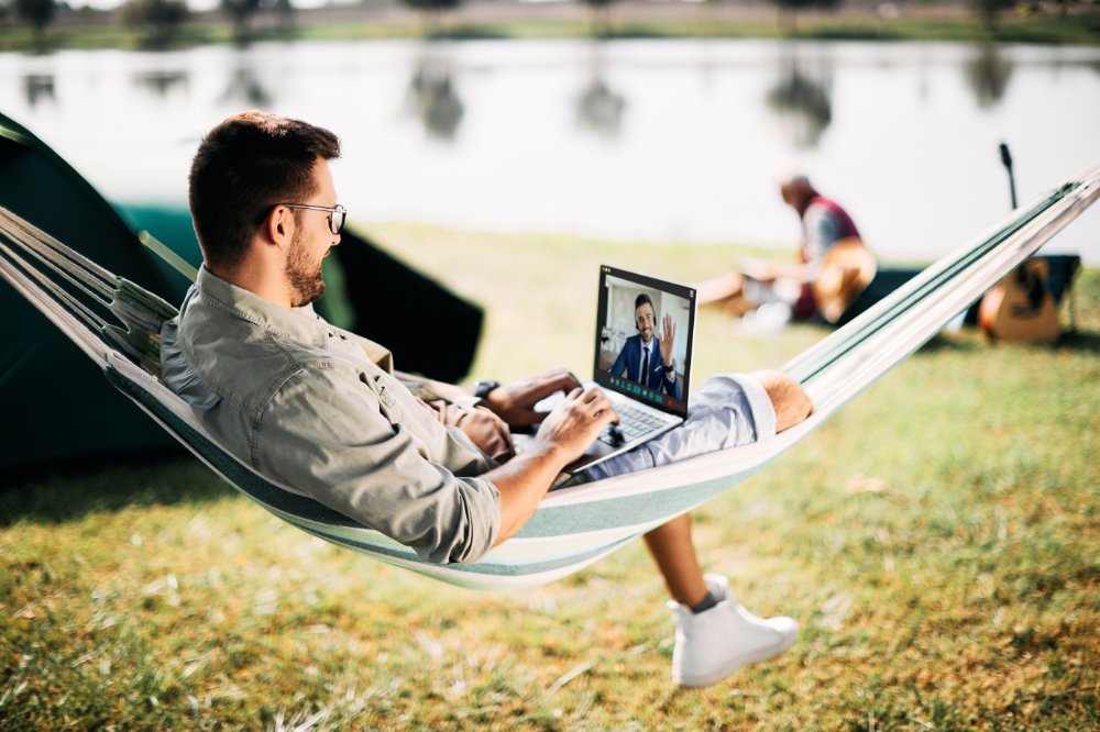 Happy entrepreneur greeting his colleague during video call while relaxing in hammock and camping in nature.