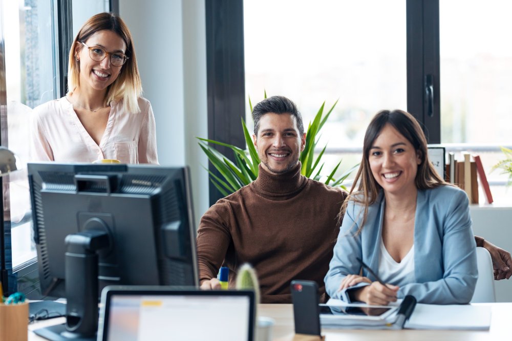 Shot of successful young business team standing around computer while looking at camera in the coworking space.