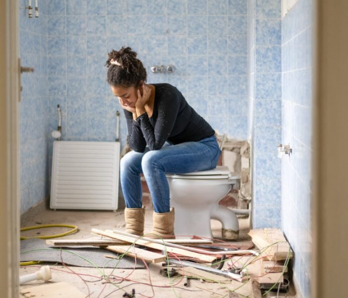 A woman takes a break on a toilet amidst a chaotic bathroom renovation.